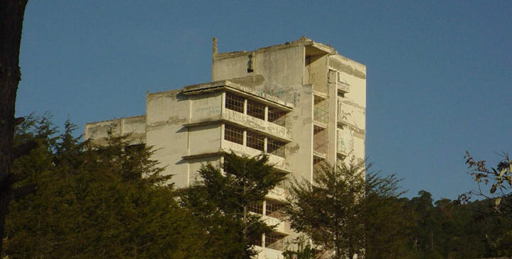 Edificio abandonado rodeado de árboles, con fachada deteriorada y paredes descascaradas, visto desde una barda de concreto bajo un cielo despejado.